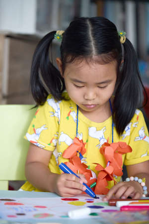 The girl happily drawing and painting on white paper. Asian girl sitting intently doing artwork on children's table.の写真素材