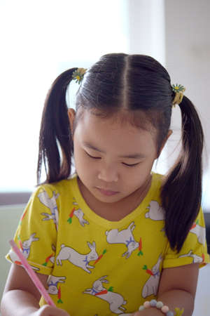 The girl happily drawing and painting on white paper. Asian girl sitting intently doing artwork on children's table.の写真素材