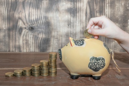 Hand puts a gold coin in a piggy bank, in front of a scale of coins sorted to growの写真素材