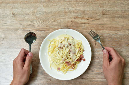 hand holding fork and spoon starving to eat spaghetti white cream sauce topping slice ham pork on...