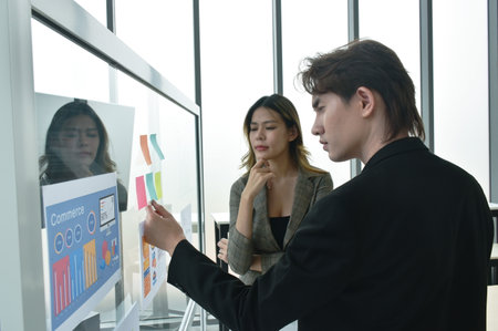 Asian business man and woman looking at paper note stick on glass board to discuss and working in...