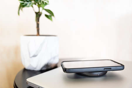 Black mobile phone on a wireless charger, with a notebook and a potted plant