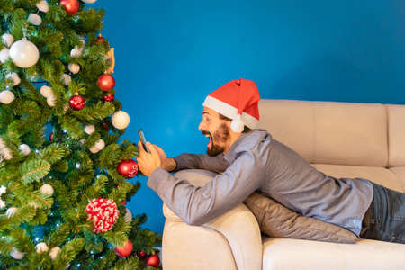Young man lying on the sofa next to decorated Christmas tree smiles with the phone