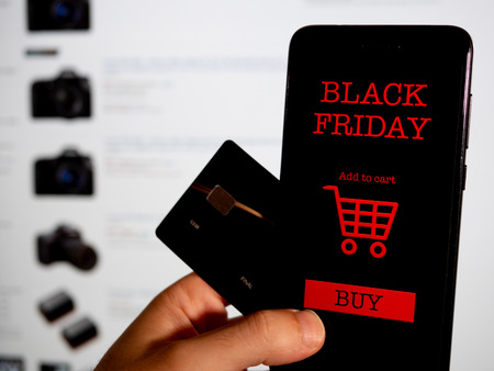 Hand holding a credit card and a phone with black friday app in front of a screen. Shopping online. First person point of viewの写真素材