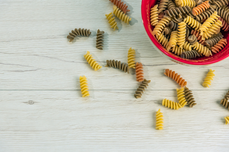 Raw uncooked three-colored Fusilli durum wheat pasta with spinach and tomato i a red bowl on white wood table, top view.の写真素材