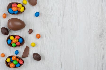 Chocolate Easter eggs and chocolate eggs filled with sweet and colorful candies, on white wood table. Copy space , top view.の写真素材