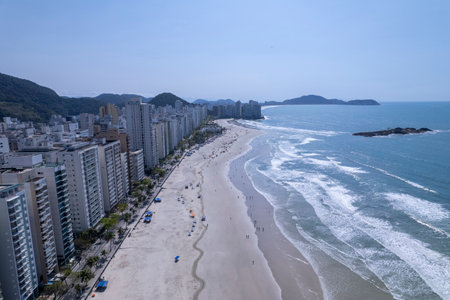This coastal landscape features Praia da Pitangueiras in Guaruja, Sao Paulo, where clear blue waters meet golden sands and tall buildings line the shore, inviting visitors to enjoy the beach.の写真素材