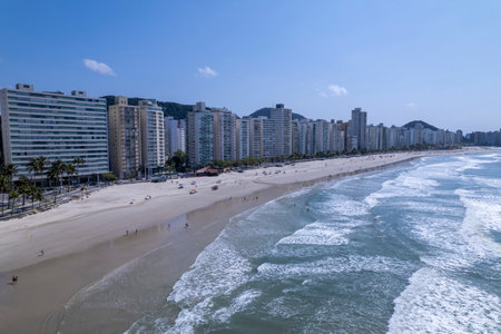 This coastal landscape features Praia da Pitangueiras in Guaruja, Sao Paulo, where clear blue waters meet golden sands and tall buildings line the shore, inviting visitors to enjoy the beach.の写真素材