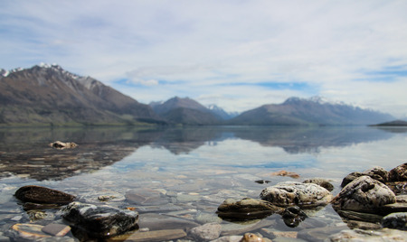 Mountains reflecting in water of Lake Wakatipu, on the way to Glenorchy, New Zealand, with rocks in the forefround.の写真素材