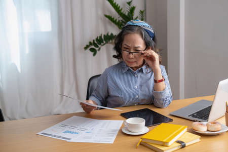 Portrait of an elderly woman working inside the house using laptops and tablets to work and check important documents for customers.の写真素材