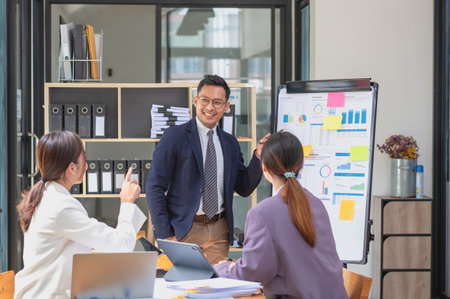 Team business three smiling and happy Asian men and women team using tablets and laptops brainstorm and plot statistics on a corporate chart board inside the company. Colleagues concept.の写真素材