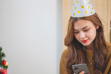 Portrait of a beautiful smiling young woman holding a smartphone on Christmas day inside the house with a green Christmas tree decorated with light bulbs and gift boxes to exchange. Christmas conceptの写真素材