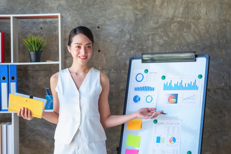 Asian businesswoman presenting business data with a board with charts, graphs hand holding a yellow notebook, And business documents lying on the table in the office, business concept.の写真素材