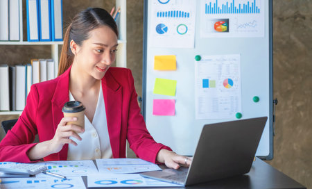 Financial, Asian businesswoman in red suit holding cup of coffee sitting on desk in office, having computer for doing accounting work at workplace to calculate annual profit by duty, Business ideaの写真素材