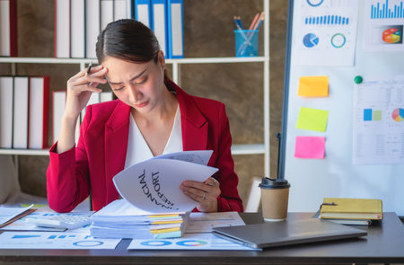 Financial, Asian businesswoman in red suit holding cup of coffee sitting on desk in office, having computer for doing accounting work at workplace to calculate annual profit by duty, Business ideaの写真素材