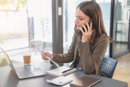 Marketing, Finance, Accounting, Planning, Businesswoman of bi nationality is talking with a customer representing a company distributor using a smartphone with laptop pen and notepad on office desk.の写真素材