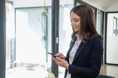 A stylish white businesswoman in a black suit standing inside the office in hands with a mobile phone, tablet, and laptop waiting for customers to contact, business people concept.の写真素材