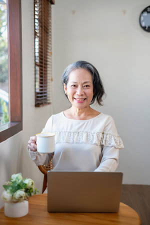 Elderly women sit and relax, stressed, watching series on laptops, reading and taking notes in notebooks and sipping tea happily inside the house with the brightest light and fresh air.の写真素材