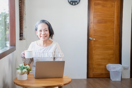 Elderly women sit and relax, stressed, watching series on laptops, reading and taking notes in notebooks and sipping tea happily inside the house with the brightest light and fresh air.の写真素材