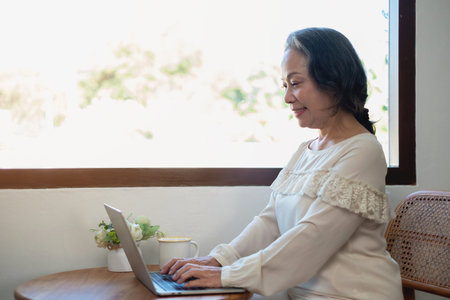 Elderly women sit and relax, stressed, watching series on laptops, reading and taking notes in notebooks and sipping tea happily inside the house with the brightest light and fresh air.の写真素材