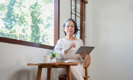 Elderly women sit and relax, stressed, watching series on laptops, reading and taking notes in notebooks and sipping tea happily inside the house with the brightest light and fresh air.の写真素材