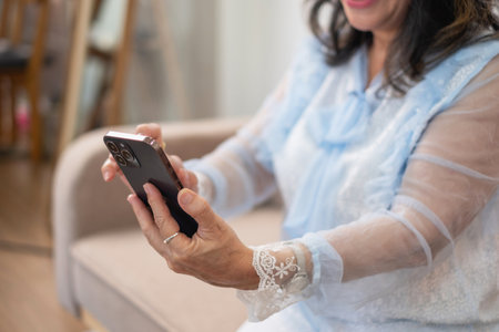 An elderly businesswoman in her hand holding a smartphone looks at the information about the company she is a senior boss inside the house and happily eats snacks and drinks coffee, Concept of WFH.の写真素材
