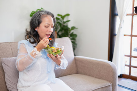 Asian senior woman sitting eating vegetable salad and healthy food and eating happily on the sofa in the house for a healthy body. healthy food conceptの写真素材