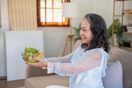 Asian senior woman sitting eating vegetable salad and healthy food and eating happily on the sofa in the house for a healthy body. healthy food conceptの写真素材