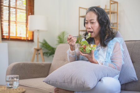 Asian senior woman sitting eating vegetable salad and healthy food and eating happily on the sofa in the house for a healthy body. healthy food conceptの写真素材