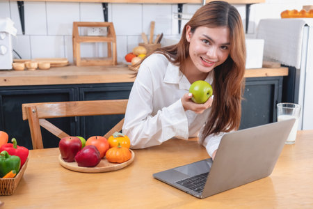 Woman with a beautiful face in a white shirt is making a healthy breakfast with bread, vegetables, fruit and milk inside the kitchen and opening her laptop for cooking lessons. healthy cooking ideas.の写真素材
