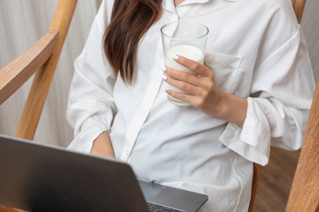 Portrait of a beautiful young woman in a white shirt holding a glass of milk and a laptop to type her own story and sitting on a wooden chair inside the house, the concept of restの写真素材