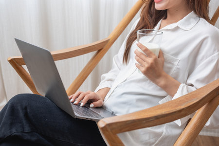 Portrait of a beautiful young woman in a white shirt holding a glass of milk and a laptop to type her own story and sitting on a wooden chair inside the house, the concept of restの写真素材