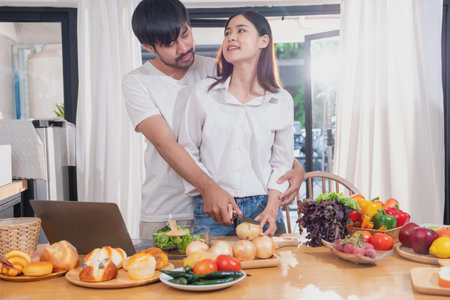 Young Asian couple cooking with fruits and vegetables and using laptop in the kitchen To cook food together within the family happily, family concept.の写真素材