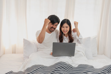 Young happy asian couple using laptop in bed watching movie, romantic time to enhance family relationship, family concept.の写真素材