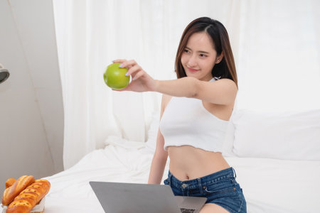 Portrait of a white Asian woman with tattoos sitting holding green apples and fruits in a healthy lifestyle. Relaxing in bed and playing on my laptop for the weekend. holiday concept.の写真素材