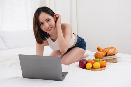 Portrait of white Asian woman with a tattoo sitting on the bed relaxing and playing laptop on weekend There was bread and fruit and red apples on a white bed to eat. holiday conceptの写真素材