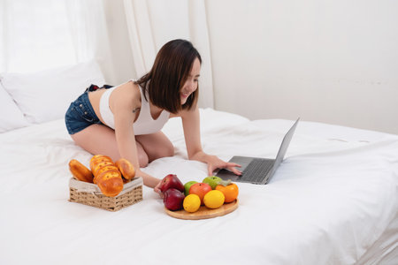 Portrait of white Asian woman with a tattoo sitting on the bed relaxing and playing laptop on weekend There was bread and fruit and red apples on a white bed to eat. holiday conceptの写真素材