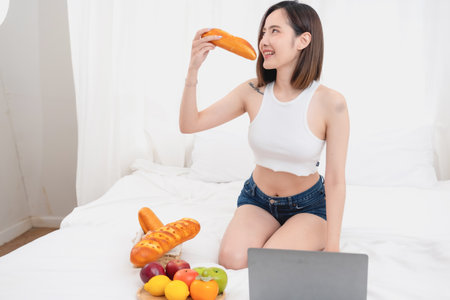 Portrait of a white Asian woman with a tattoo sitting and holding bread and fruit lying in bed eating happily. Relax in bed and play on your laptop on the weekends. holiday concept.の写真素材