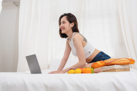 Portrait of white Asian woman with a tattoo sitting on the bed relaxing and playing laptop on weekend There was bread and fruit and red apples on a white bed to eat. holiday conceptの写真素材