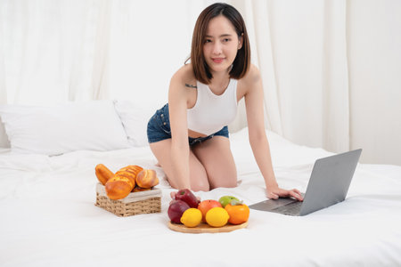 Portrait of white Asian woman with a tattoo sitting on the bed relaxing and playing laptop on weekend There was bread and fruit and red apples on a white bed to eat. holiday conceptの写真素材