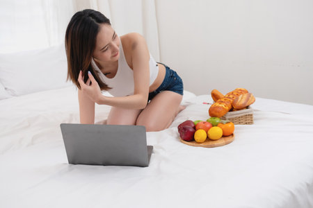 Portrait of white Asian woman with a tattoo sitting on the bed relaxing and playing laptop on weekend There was bread and fruit and red apples on a white bed to eat. holiday conceptの写真素材