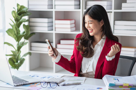 Happy Asian businesswoman in red suit showing happy gesture holding smartphone at desk Portrait of beautiful smiling businesswoman working at a modern workstation.の写真素材