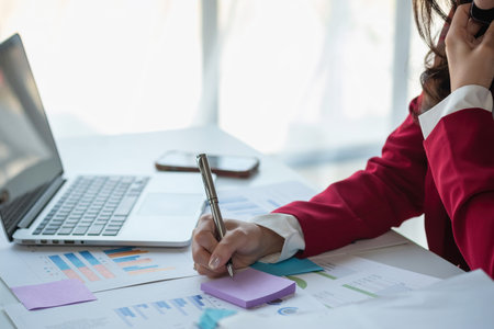 Asian businesswoman in red suit calculating company sales with calculator, laptop, and tablet on table Interior of businesswoman office at the modern workstation.の写真素材