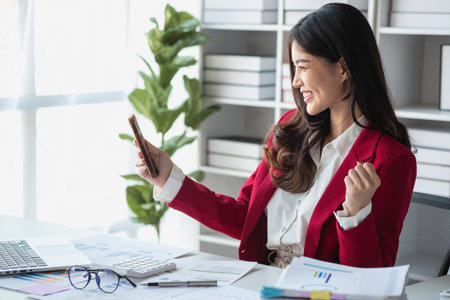 Happy Asian businesswoman in red suit showing happy gesture holding smartphone at desk Portrait of beautiful smiling businesswoman working at a modern workstation.の写真素材
