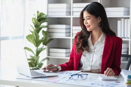 Asian businesswoman in red suit calculating company sales with calculator, laptop, and tablet on table Interior of businesswoman office at the modern workstation.の写真素材