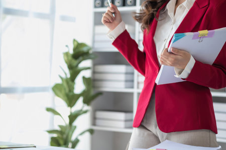 Asian businesswoman in red suit calculating company sales with calculator, laptop, and tablet on table Interior of businesswoman office at the modern workstation.の写真素材