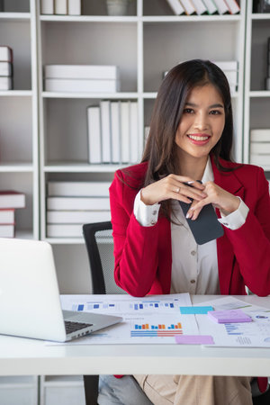 Asian businesswoman in red suit calculating company sales with calculator, laptop, and tablet on table Interior of businesswoman office at the modern workstation.の写真素材
