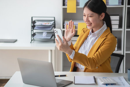Young Asian businesswoman in a yellow suit showing joy through a laptop and smartphone to colleagues for accomplishing a company project with set objectives, the concept of success.の写真素材