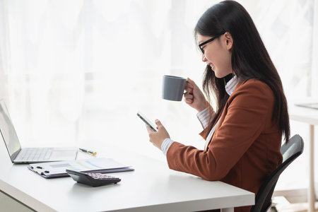Young businesswoman with cheerful face sitting drinking coffee and using laptop in office Check email on the computer, sitting at a desk. Interior of the modern office.の写真素材