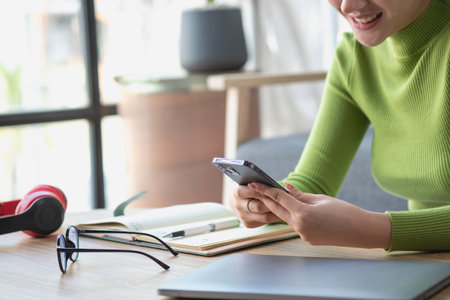 Asian woman with a beautiful face in a green long-sleeve shirt sitting and relaxing watching movies Listening to music on a laptop, mobile phone, and headphones happily inside living room at home.の写真素材